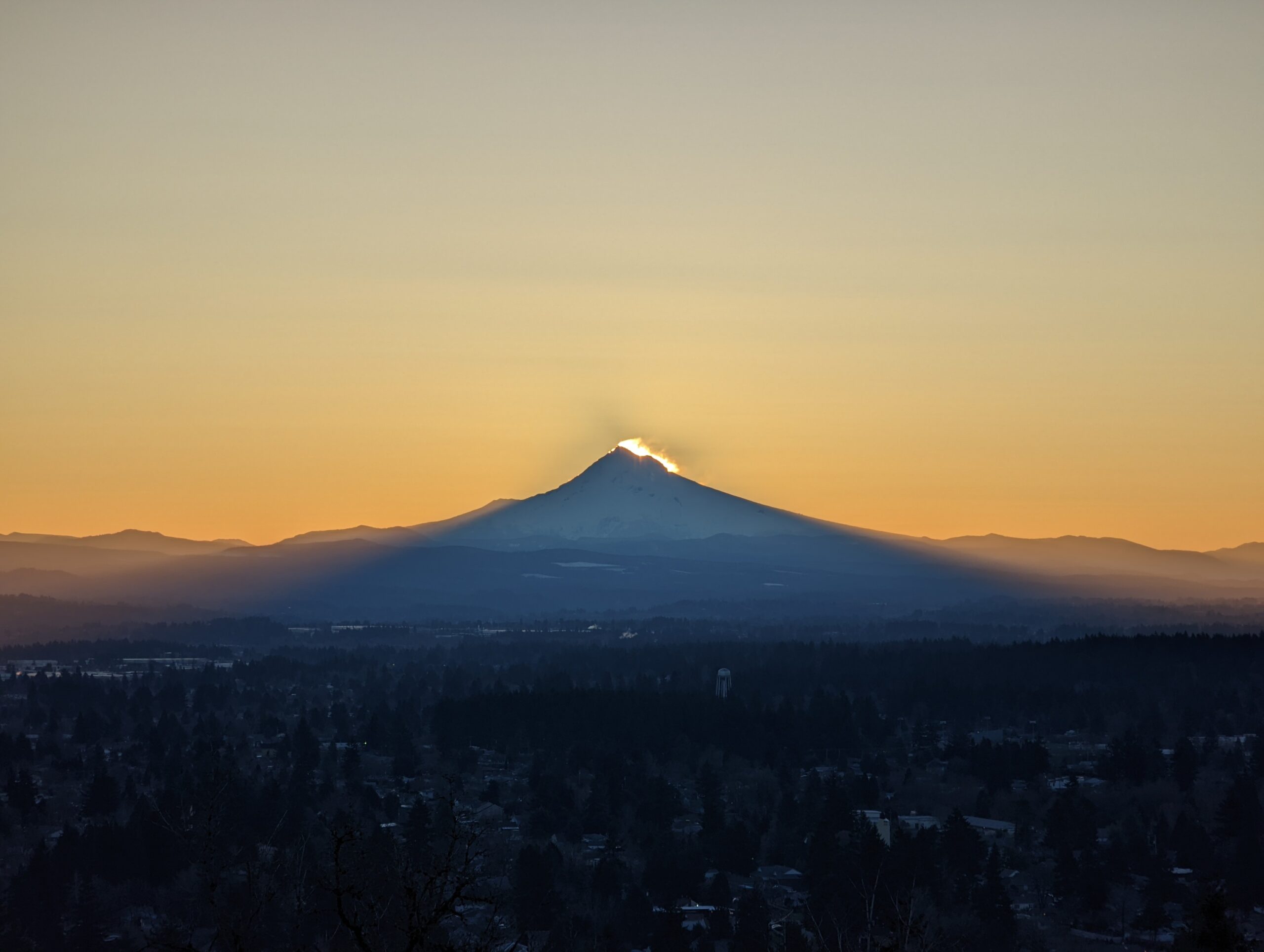 Mount Hood as seen from Rocky Butte before sunrise. The sun is starting to appear in the wind-blown snow from the summit, and the mountain's shadow is being cast on the city.