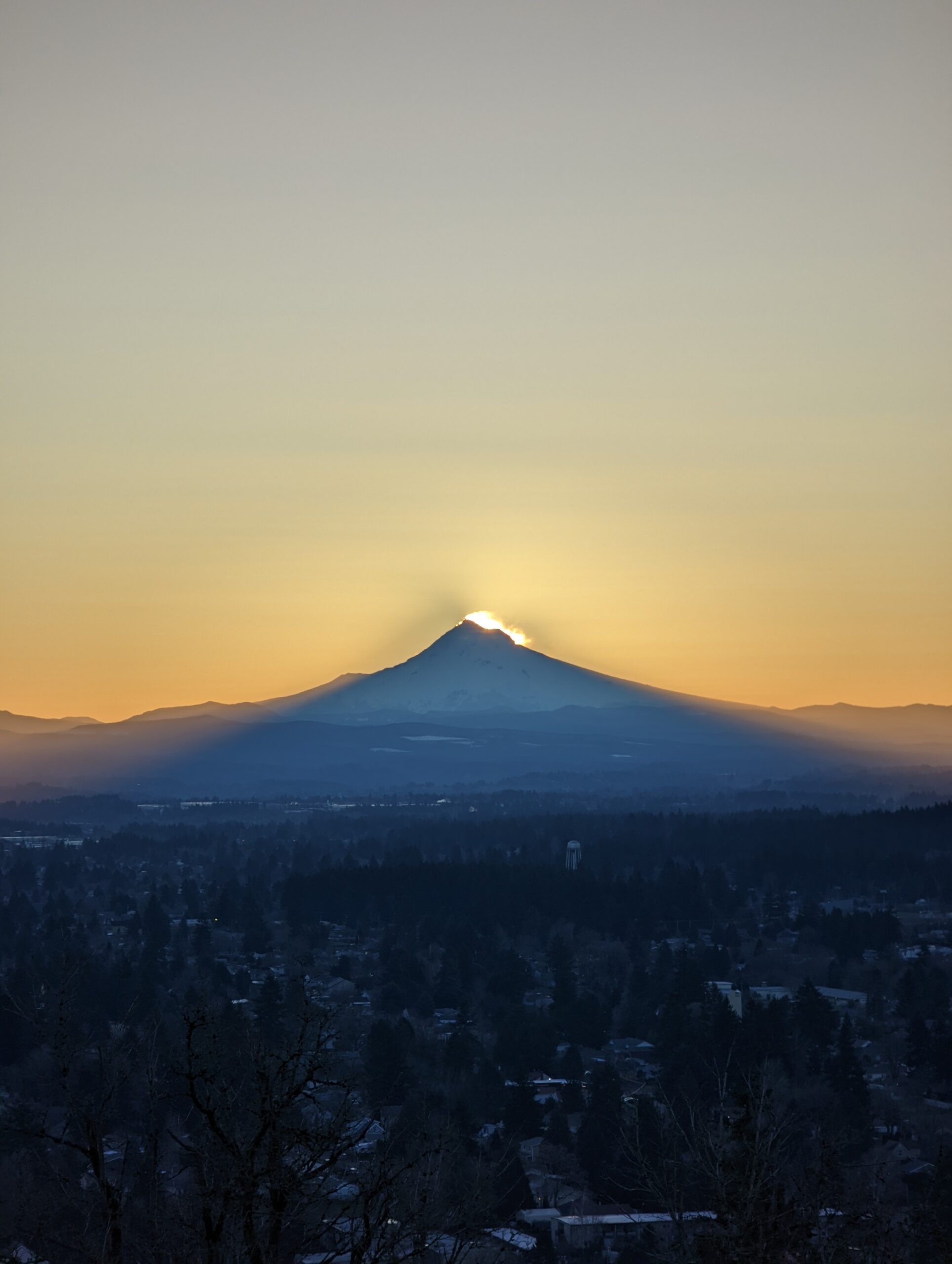 Mount Hood as seen from Rocky Butte before sunrise. The sun is starting to appear in the wind-blown snow from the summit, and the mountain's shadow is being cast on the city.